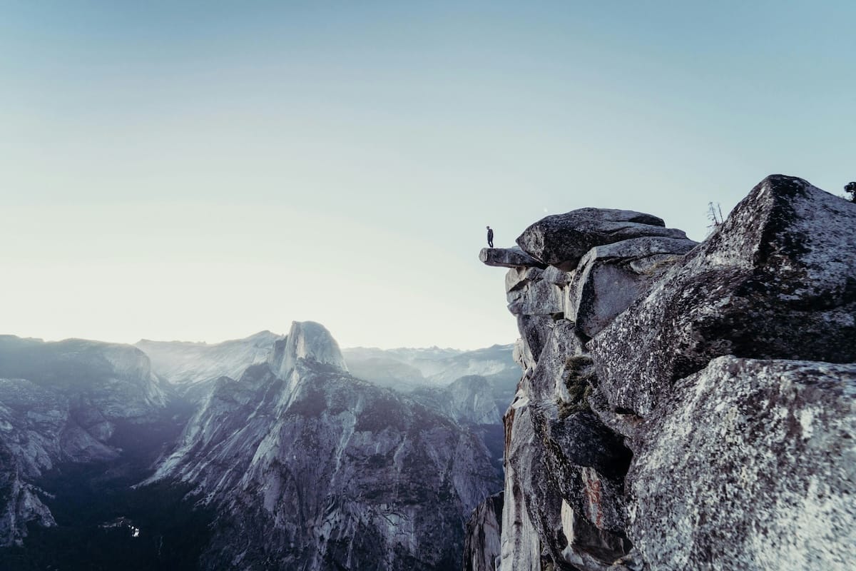 Man standing on a small ledge on the side of a mountain overlooking a mountain range