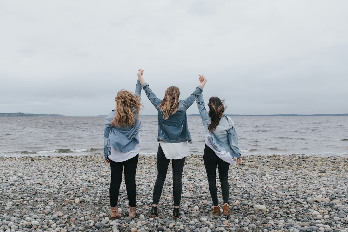 Women holding hands in the air while standing on a beach facing the water