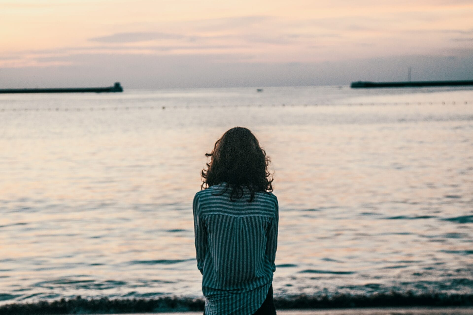 Woman standing facing the open water while standing on a beach at sunset