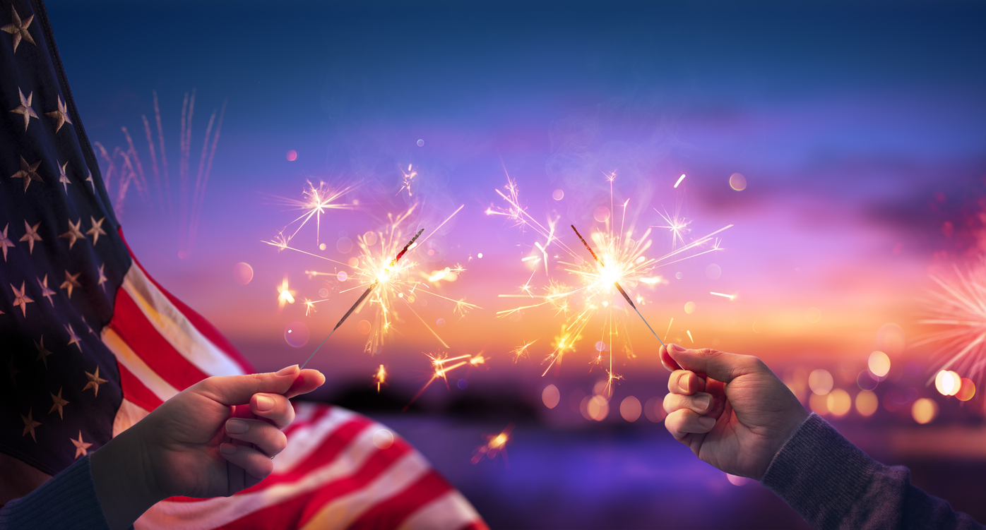 USA-Celebration-With-Hands-Holding-Sparklers-And-American-Flag-At-Sunset-With-Fireworks