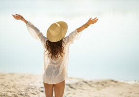 Romantic woman rising hands feeling free, relaxing on a beach against a beautiful sea bokeh background. Tanned girl in fashionable trendy boho style rear view. Travel vacation, summer outdoor pleasure concept.