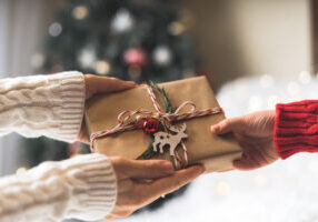 Woman in sweater giving a wrapped Christmas gift box to child. Glowing snow bokeh, fir tree. Winter holidays.