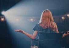 Pan left view of young woman smiling and gesticulating while sitting on wheelchair in spotlight and talking to audience during performance in dark theater